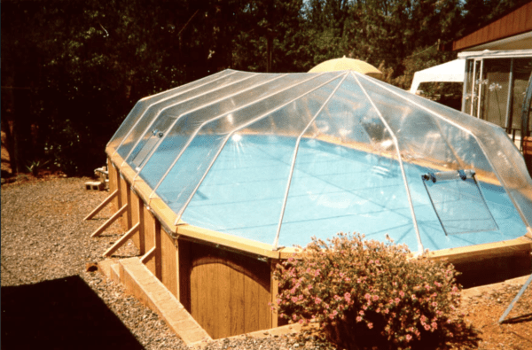 A Round Above Ground Sun Dome cover over an octagonal wooden pool deck, with a residential building and shrubbery in the background.